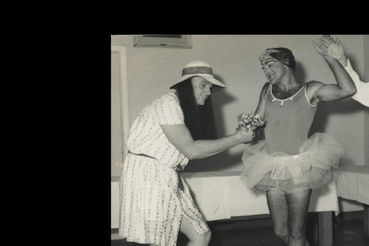 Two male dancers, one in a leotard and tutu, pose with an actor in drag who is handing them a bouquet of flowers. 
