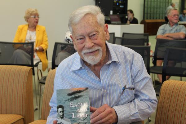 Senior gentleman wearing button up long sleeve blue shirt holding a book and smiling at the camera