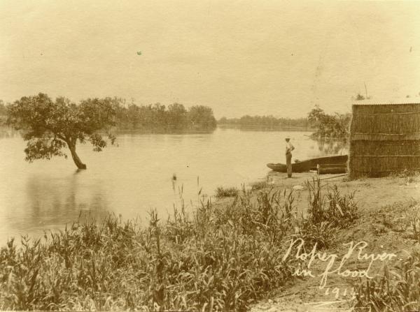 Sepia colour image of man standing on bank and looking at the Roper River with a stack of hay bales behind him