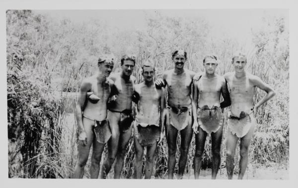 Six men standing shoulder to shoulder, wearing only lily-pad “loin cloths” and lilies in their hair.