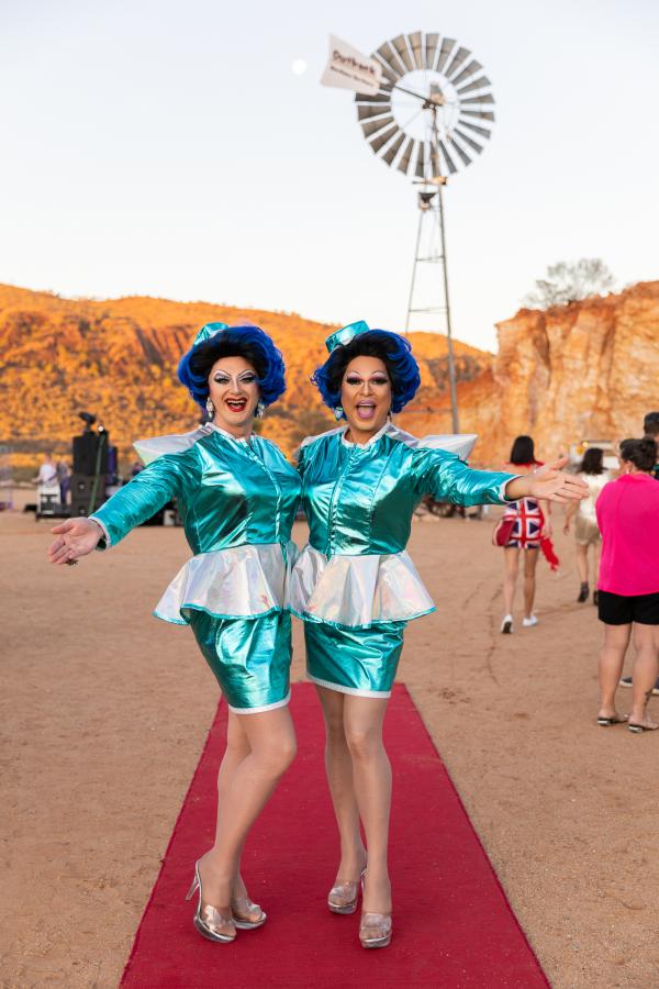 Two well-dressed and welcoming “hostesses” in short satin skirts on the red carpet in front of a windmill and desert range.